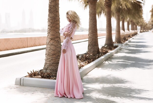 Rubia bien formada junto al mar en Dubai, palmeras, vestidos hermosos y calientes, sesión de fotos de moda de estilo de vida soleado de verano, vestido ondeando en el viento, tranquilo y relajado cerca de la piscina, peinado, maquillaje