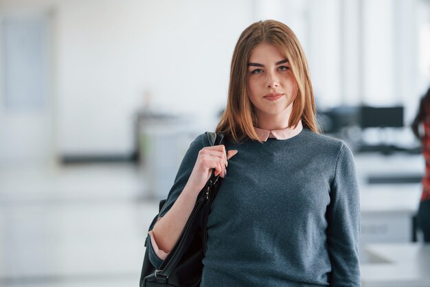 Rubia con bolso negro en manos. Retrato de mujer joven atractiva de pie en la oficina