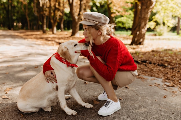 Rubia fascinante con adorable labrador pasando el día juntos en el parque de otoño. Conmovedora foto de niña en ropa de temporada abrazando a su amado perro.