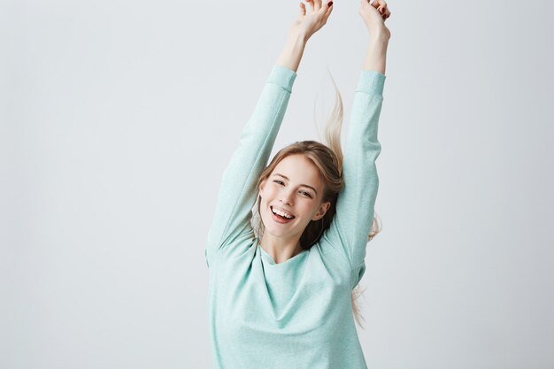 Rubia joven y bella mujer en azul claro superior estirando los brazos en alegre estado de ánimo como celebrar la victoria. Ampliamente sonriente mujer mostrando dientes blancos y emociones positivas, divirtiéndose en el interior.