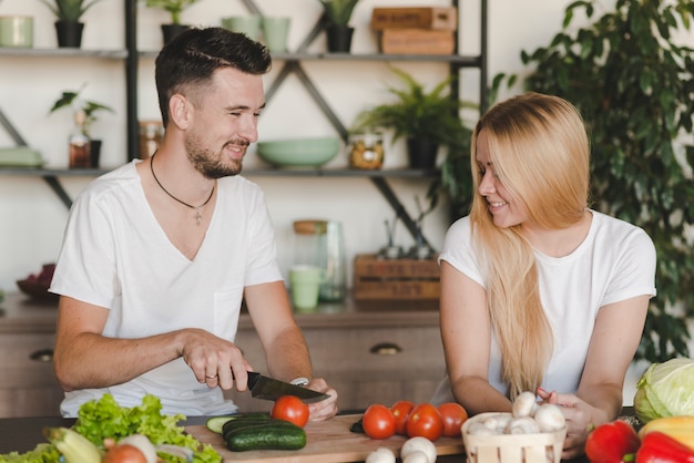 Rubia joven mirando hombre cortando verduras con cuchillo