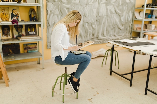 Rubia joven sentada en el bosquejo de pintura de taburete en el taller