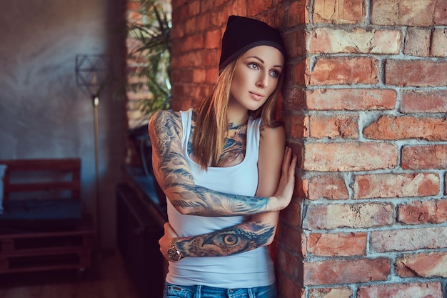 Una rubia sexy tatuada con una camiseta y un sombrero posando contra una pared de ladrillos.