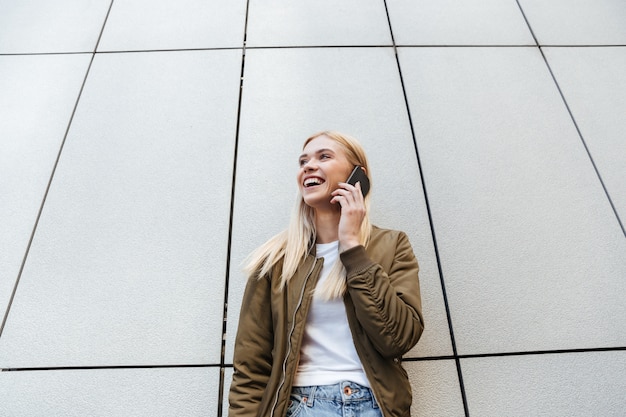 Rubia sonriente hablando por teléfono mientras está parado cerca de la pared