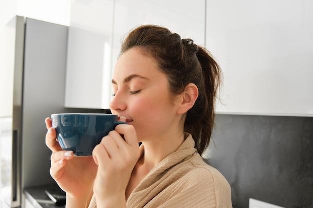 Rutina diaria y estilo de vida joven hermosa mujer en bata de baño parada en la cocina con una taza de café