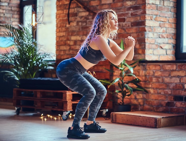 Rutina de piernas para mujer. Mujer deportiva rubia haciendo sentadillas en una habitación con interior de loft.