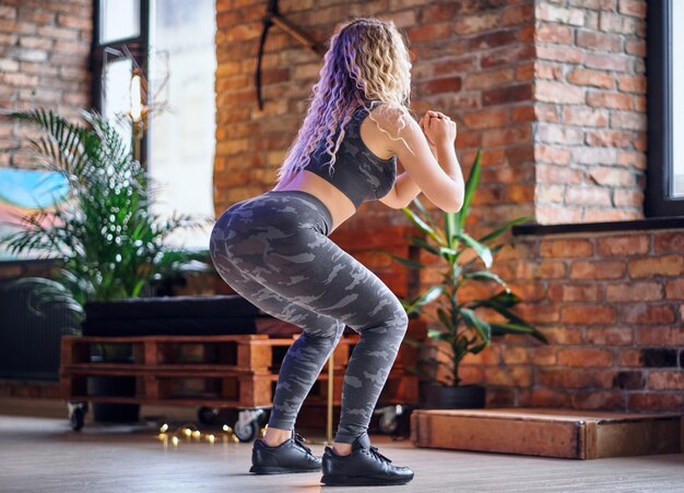 Rutina de piernas para mujer. Mujer deportiva rubia haciendo sentadillas en una habitación con interior de loft.