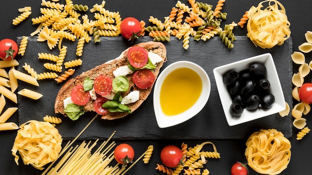 Sabrosa comida italiana con pasta cruda; Aceitunas negras y cuenco de aceite sobre superficie negra.