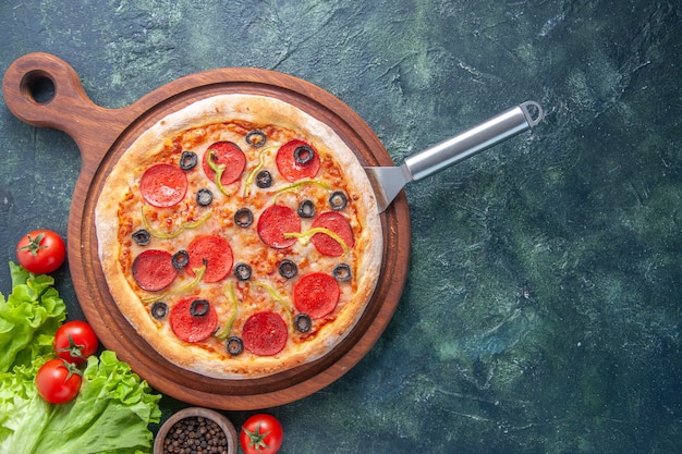 Sabrosa pizza casera sobre tabla de madera, botella de aceite, tomates, pimienta, paquete verde sobre superficie oscura