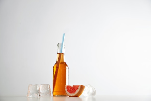 Sabrosa sidra fresca en botella transparente con pajita azul cerca de cubitos de hielo y rodaja de pomelo rojo aislado en blanco