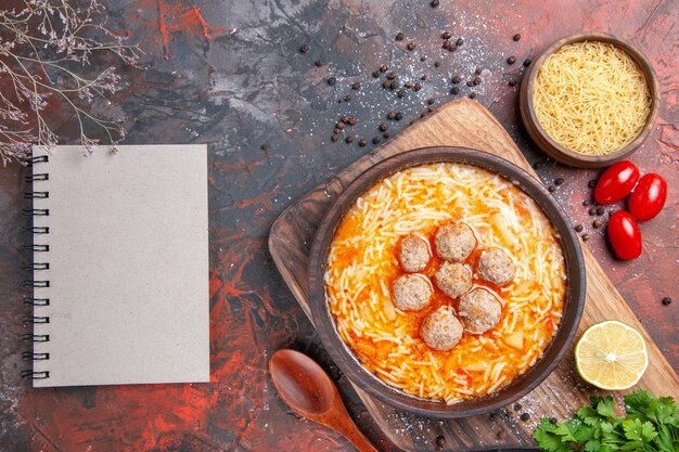 Sabrosa sopa de albóndigas con fideos a bordo de pasta de madera de limón un manojo de tomates verdes pimiento y cuaderno sobre mesa oscura