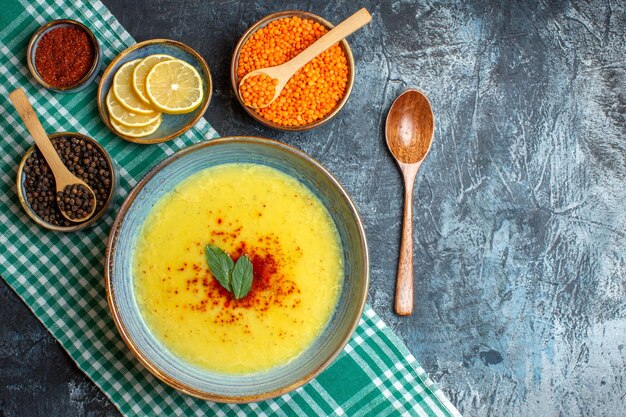 Sabrosa sopa servida con menta y pimienta en una toalla despojada verde junto a una cuchara de madera de limón picado y diferentes especias de guisantes amarillos sobre fondo azul.