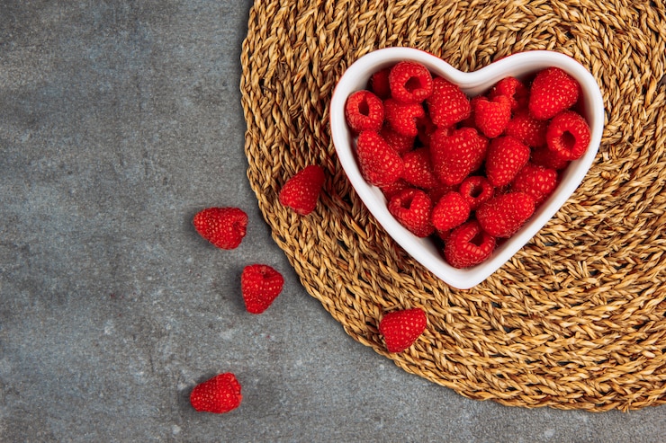 Foto gratuito sabrosas frambuesas en un plato en forma de corazón sobre mantel de mimbre y fondo de asfalto gris, plano.