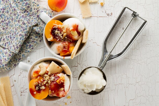 Sabroso helado de vainilla apetitoso fresco con nueces, albaricoques, waffles y jarabe en la mesa blanca con ingredientes para hacer el postre. Vista superior.