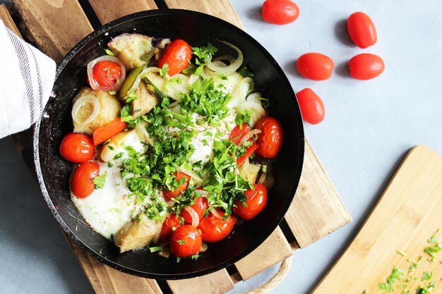 Sabrosos huevos fritos con tomates cherry, patatas y cebollas en una sartén sobre un soporte de madera sobre la mesa