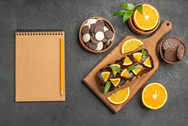 Sabrosos pasteles cortan naranjas con galletas junto al cuaderno en la tabla de cortar en la mesa oscura