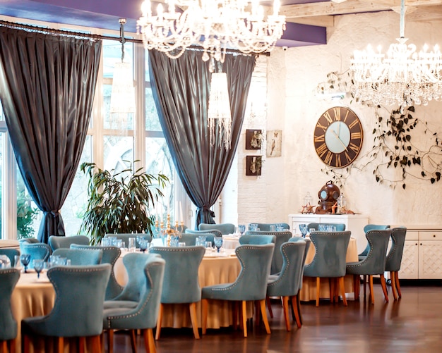 Sala del restaurante con sillas turquesas, paredes blancas, ventanas francesas y cortinas.