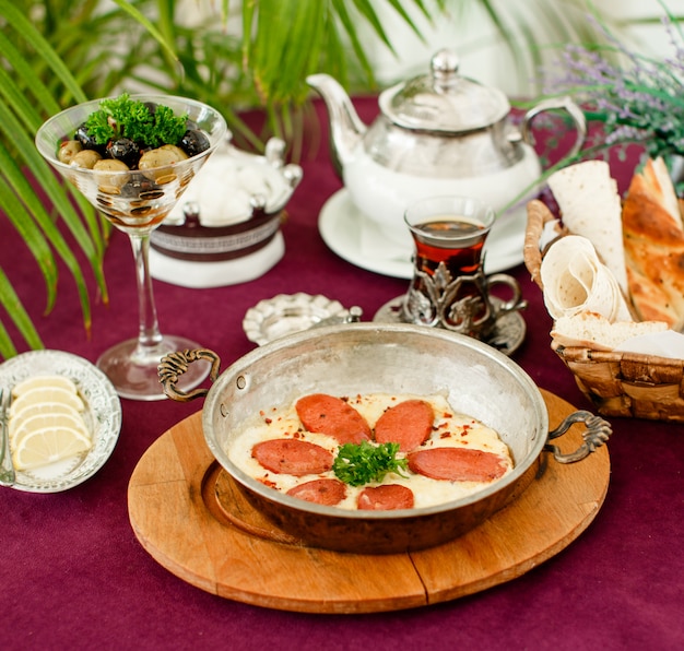 Foto gratuita salchicha turca con huevos en sartén de acero, tetera, aceitunas y pan