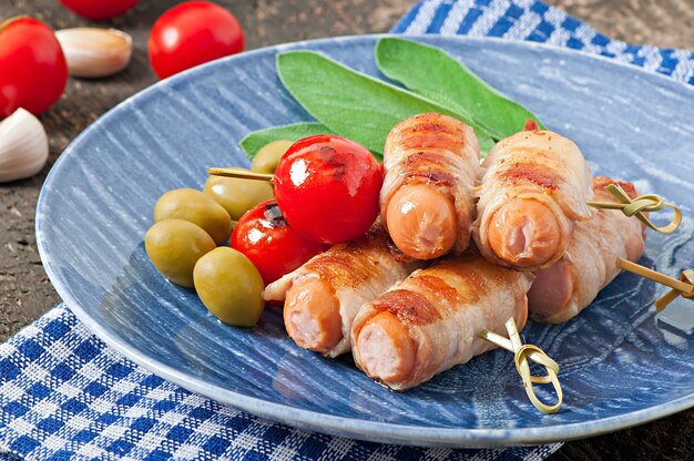 Salchichas a la parrilla envueltas en tiras de tocino con tomates y hojas de salvia