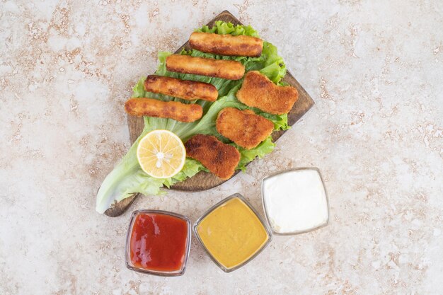 Salchichas a la plancha con nuggets de pollo sobre un trozo de lechuga sobre una tabla de madera con salsas a un lado.