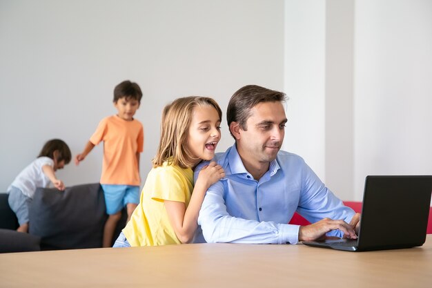 Salió de la hermosa niña mirando la pantalla del portátil y abrazando a papá. Padre de mediana edad caucásico que trabaja en casa cuando niños lindos jugando en el sofá. Concepto de infancia, paternidad y tecnología digital.