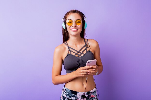 Salió mujer deportiva con sincera sonrisa posando en ropa deportiva con estilo en estudio. Usando teléfonos inteligentes y escuchando música.