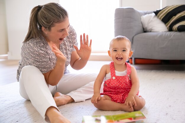 Salió a la niña sentada en el suelo y jugando con la madre. Mamá rubia alegre divirtiéndose con su hermosa hija, aplaudiendo y gritando algo. Concepto de familia, maternidad y estar en casa