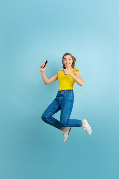 Saltando alto con el teléfono inteligente. Retrato de muchacha adolescente caucásica en azul