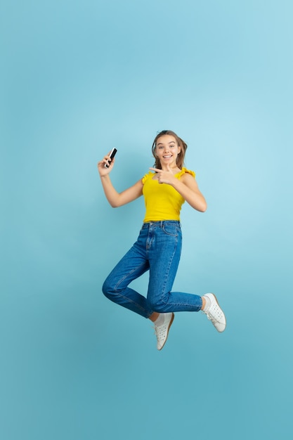 Saltando alto con el teléfono inteligente. Retrato de muchacha adolescente caucásica en azul