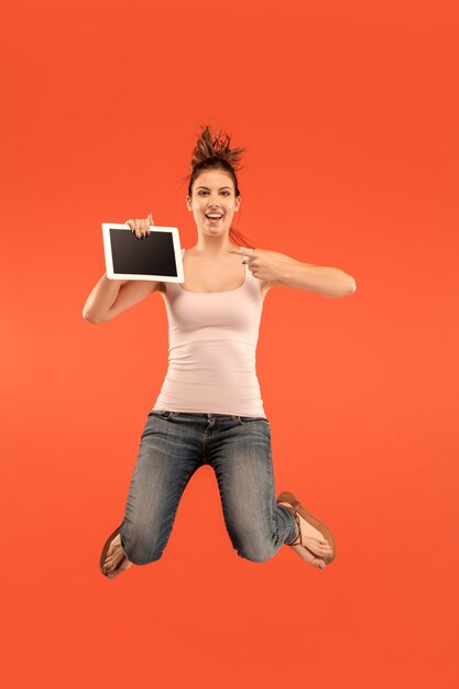 Salto de mujer joven sobre fondo azul de estudio con gadget portátil o tableta mientras salta. Chica corriendo en movimiento o movimiento.