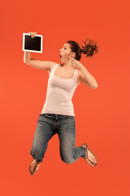 Salto de mujer joven sobre fondo azul de estudio con gadget portátil o tableta mientras salta. Chica corriendo en movimiento o movimiento.