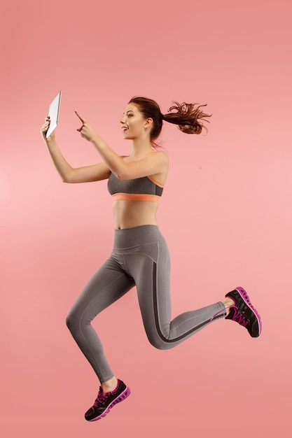 Salto de mujer joven sobre fondo azul de estudio con gadget portátil o tableta mientras salta. Chica corriendo en movimiento o movimiento.