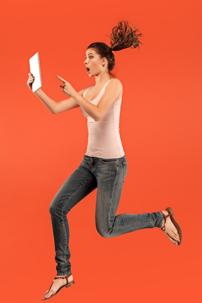 Salto de mujer joven sobre fondo azul de estudio con gadget portátil o tableta mientras salta. Chica corriente en movimiento o movimiento.