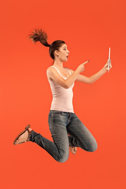 Salto de mujer joven sobre fondo azul de estudio usando un dispositivo portátil o tableta mientras salta. Chica corriendo en movimiento o movimiento. Concepto de expresiones faciales y emociones humanas. Gadget en la vida moderna