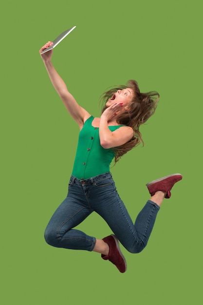 Salto de mujer joven sobre fondo verde de estudio usando un gadget portátil o tableta mientras salta. Chica corriendo en movimiento o movimiento. Concepto de expresiones faciales y emociones humanas. Gadget en la vida moderna