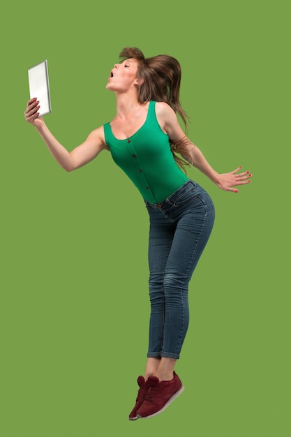 Salto de mujer joven sobre fondo verde de estudio usando un gadget portátil o tableta mientras salta.