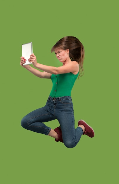 Salto de mujer joven sobre fondo verde de estudio usando un gadget portátil o tableta mientras salta.