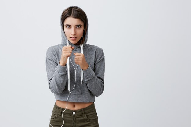 Salud y Belleza. Ciérrese para arriba de muchacha caucásica apuesta atractiva joven con el pelo largo oscuro en sudadera con capucha gris deportiva que lleva a cabo las manos en actitud de la lucha, entrenando para la competencia de la caja en gimnasio.