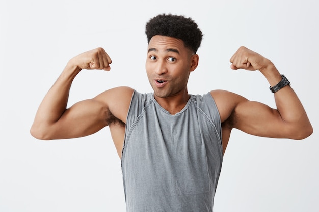 Salud y Belleza. Retrato de hombre africano maduro atractivo de piel negra con cabello rizado en camisa gris deportiva que muestra los músculos del brazo, mirando a la cámara con expresión de la cara emocionada.