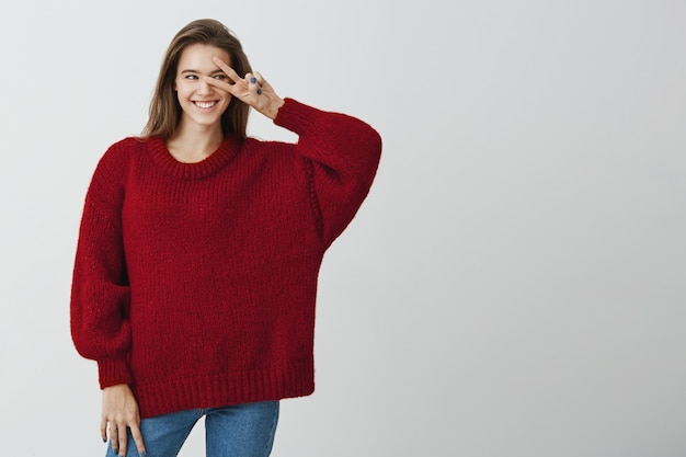 Saluda a nuevas oportunidades por delante. Foto de estudio de mujer feliz guapa en suéter holgado de moda con signo v sobre ojo y sonriendo ampliamente, de humor juguetón y coqueteo