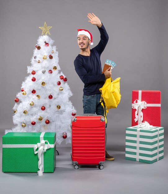 saludando a joven con gorro de Papá Noel y maleta roja comprobando su mochila amarilla alrededor de diferentes regalos
