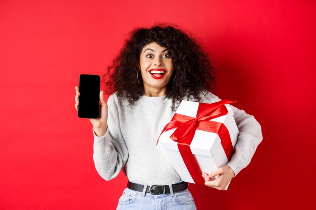 San Valentín y día de los enamorados. Mujer sonriente emocionada con cabello oscuro y rizado, mostrando la pantalla vacía del teléfono inteligente y sosteniendo un regalo sorpresa en vacaciones, mostrando promoción en línea, fondo rojo