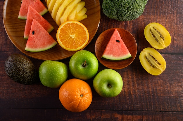 Foto gratuita sandía, naranja, piña, kiwi cortado en rodajas con manzanas y brócoli en un plato de madera y mesa de madera.