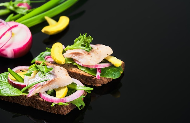 Sándwich abierto con pan de centeno oscuro, arenque matias marinado, pepino encurtido y cebolla roja en la mesa de espejo oscuro. Smorrebrod danés, enfoque selectivo y espacio de copia