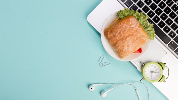 Sandwich, reloj despertador, auriculares en la computadora portátil contra el fondo azul