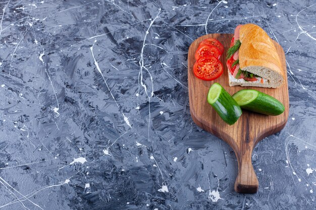 Sándwich, rodajas de pepino y tomates en tabla de cortar en azul.