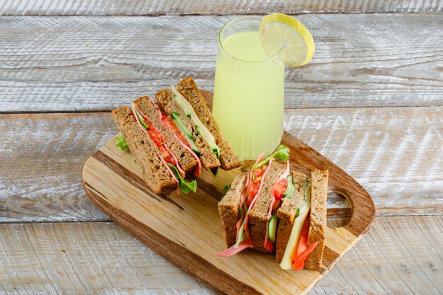 Sándwich de vegetales con queso, jamón, limonada en madera y tabla de cortar, vista de ángulo alto.