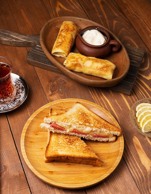 Sándwiches de club con salami, tocino y blinchik servidos con yogurt en un plato de madera con té