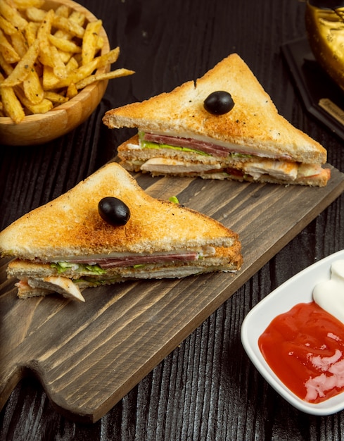 Sándwiches de club con salami, tocino y papas fritas servidos con salsa de tomate, mayonesa en un plato de madera.
