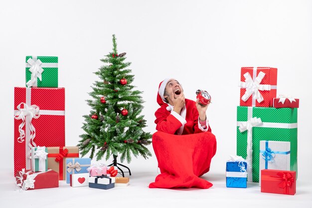 Santa claus mirando arriba sentado en el suelo y mostrando el reloj cerca de regalos y árbol de Navidad decorado